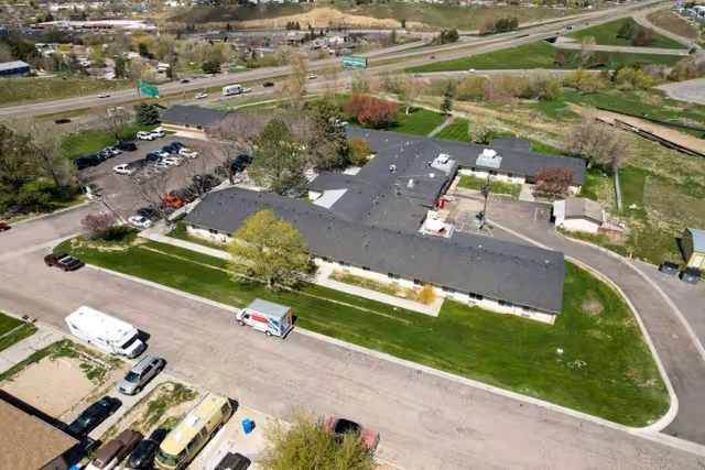 Aerial view of Monte Vista Hills Health Care Center showing a single-story building with a dark roof surrounded by green lawns, trees, and parking areas. Nearby roads and a highway with vehicles are visible in the background.