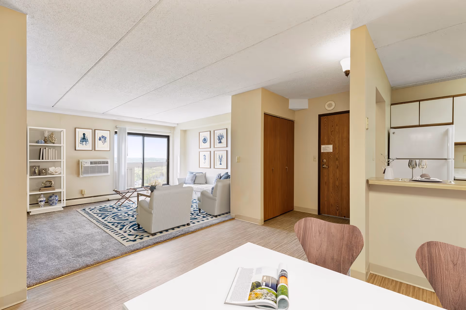 Interior view of a senior living apartment at Pathways On the Park showing a living room with two armchairs, a sofa, a patterned rug, wall art with sea creatures, a bookshelf with decorative items, and a sliding glass door leading to a balcony. Adjacent to the living room is a dining area with a white table and wooden chairs, and a kitchen with a refrigerator and countertop with two wine glasses and a small vase.