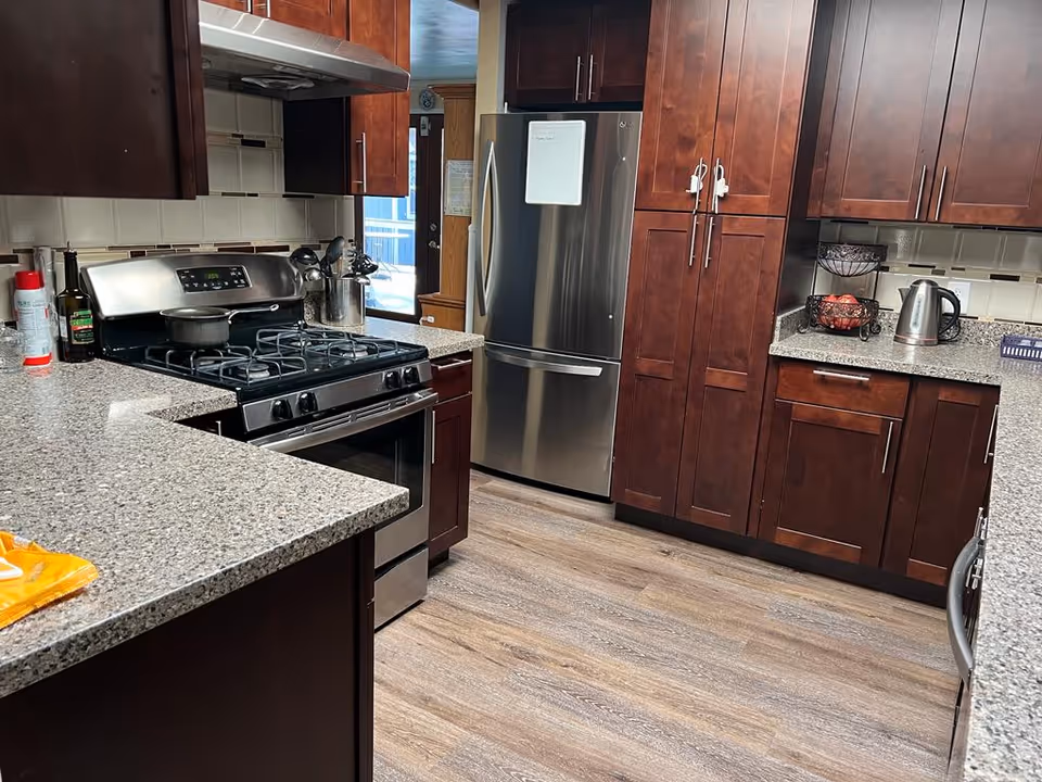 Modern kitchen with dark wood cabinets, granite countertops, a stainless steel gas stove with pots on it, a stainless steel refrigerator, and various kitchen items including a kettle and fruit basket on the counter.