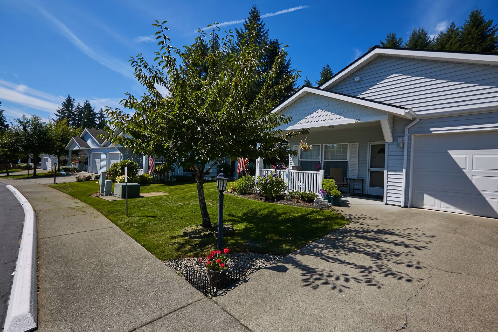 Exterior view of a single-story residential building with a small porch, garage, and well-maintained lawn with a tree and flowers under a clear blue sky.