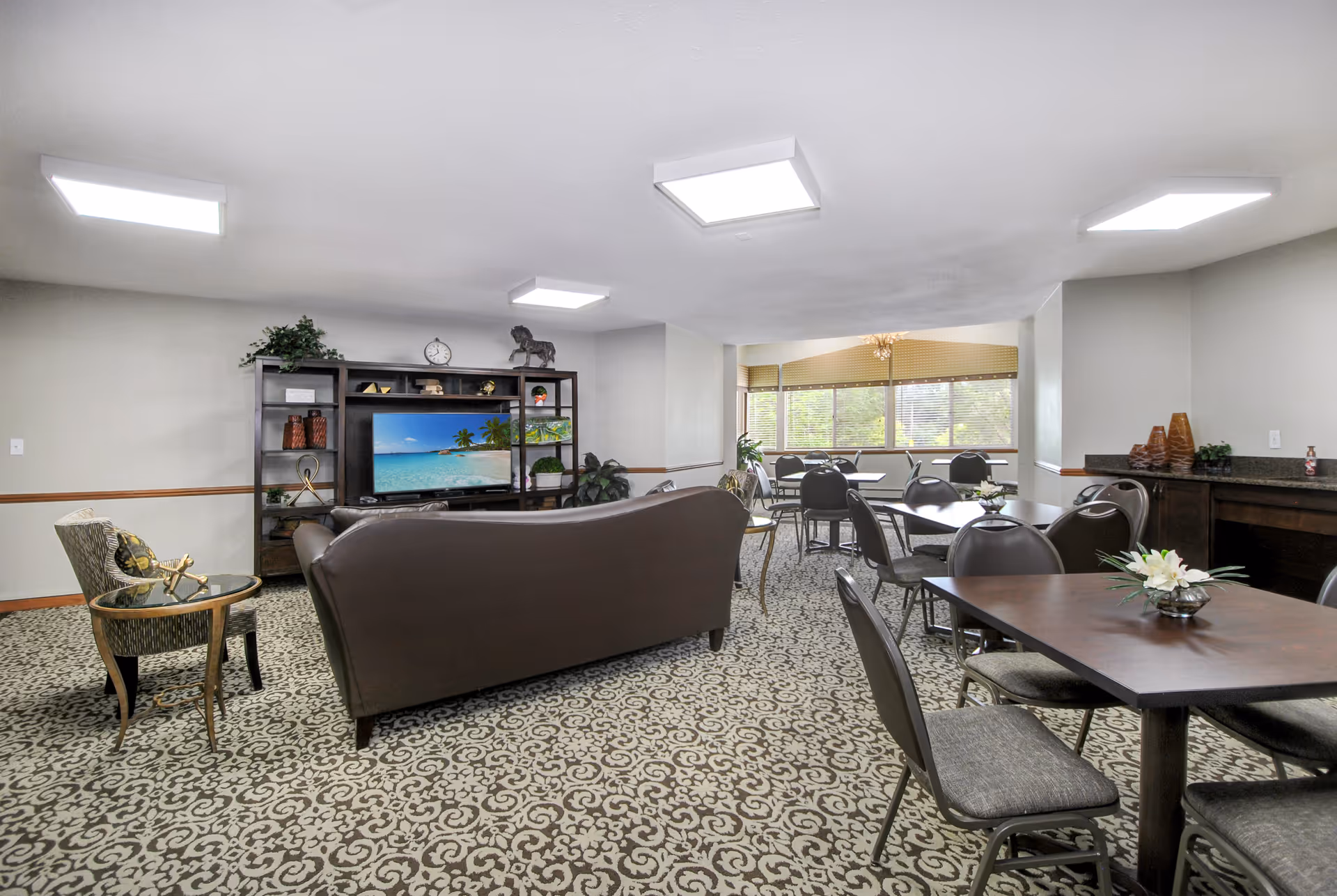A spacious common area with patterned carpet, featuring a brown leather sofa facing a large entertainment center with a TV displaying a beach scene. Several tables with chairs are arranged near large windows letting in natural light. Decorative plants and vases are placed around the room.