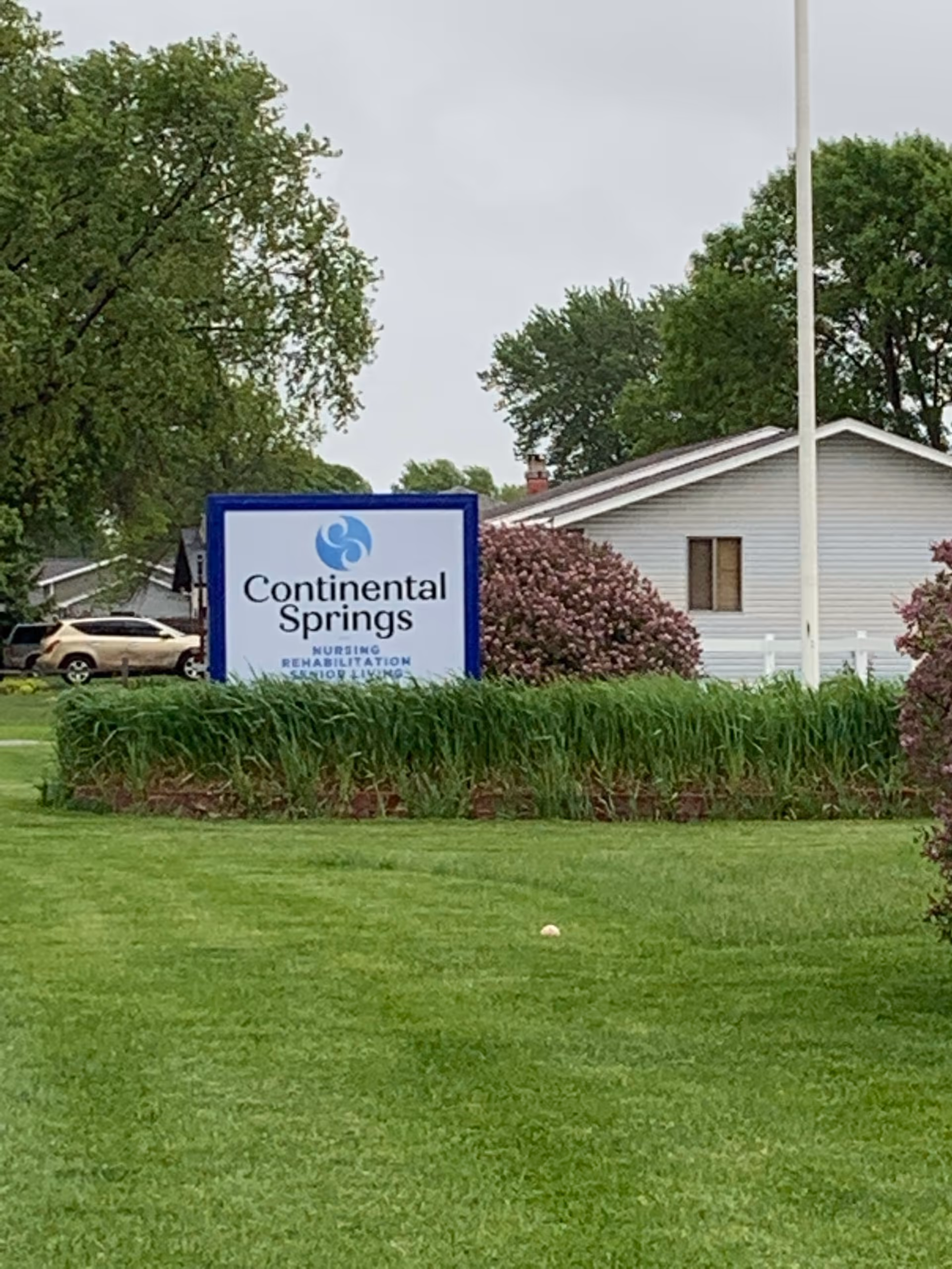 Outdoor view of a nursing and senior living facility sign that reads 'Continental Springs Nursing Rehabilitation Senior Living' surrounded by green grass, bushes, and trees with a house and parked cars in the background under an overcast sky.