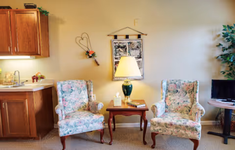 Small living area with two floral upholstered armchairs flanking a wooden side table and lamp, a kitchenette on the left and a TV on the right.