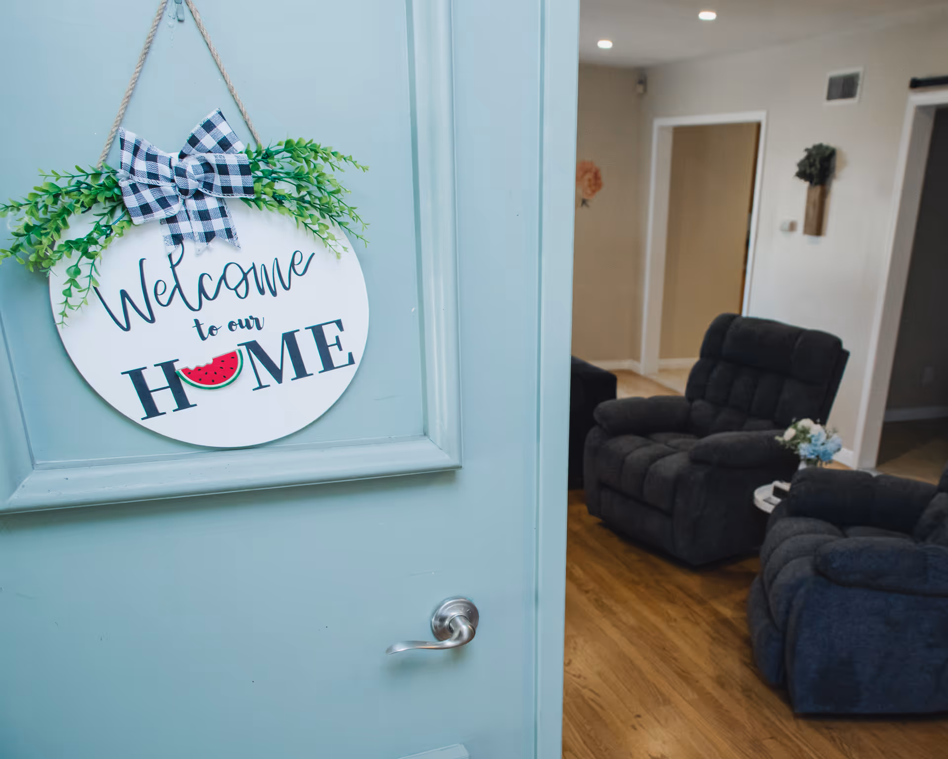 Partially open teal door with a 'Welcome to our HOME' sign revealing a living room with gray recliner chairs and hardwood floors.