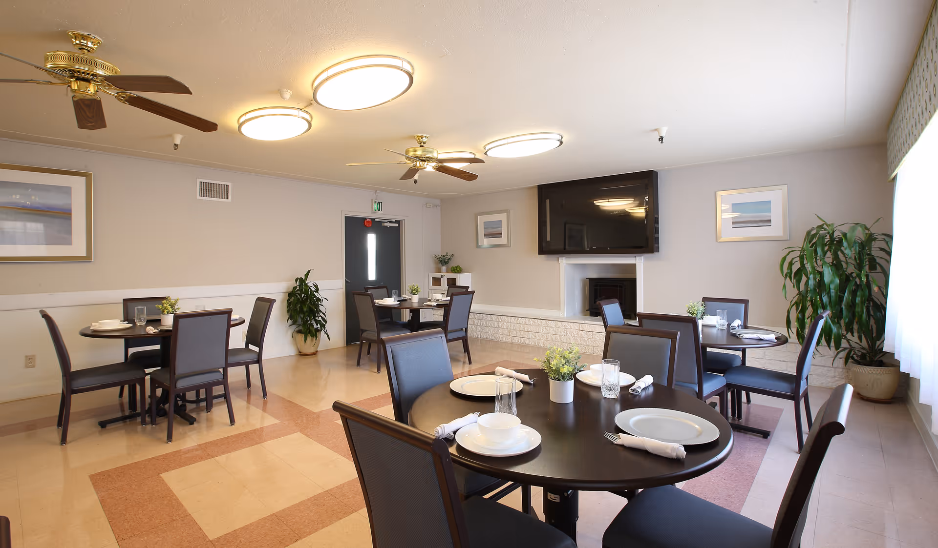 Bright dining room with round tables set for meals, chairs, ceiling fans, a wall-mounted TV above a fireplace, and potted plants.