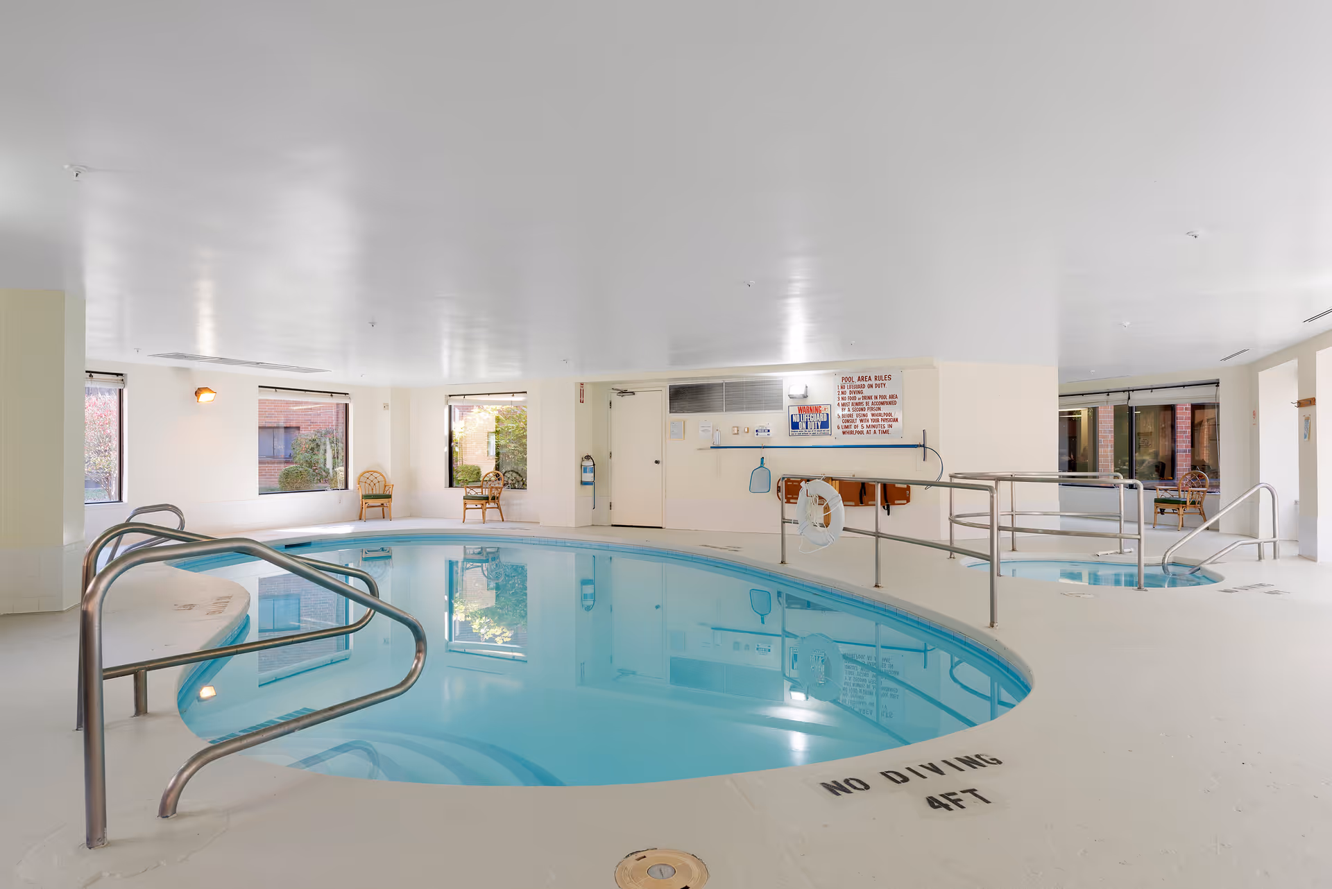 Indoor swimming pool area with a small hot tub adjacent to it. The pool has metal handrails and steps leading into the water. There are large windows letting in natural light, a few chairs placed near the windows, and safety signs on the wall. The floor around the pool is white with 'NO DIVING 4FT' marked near the edge.