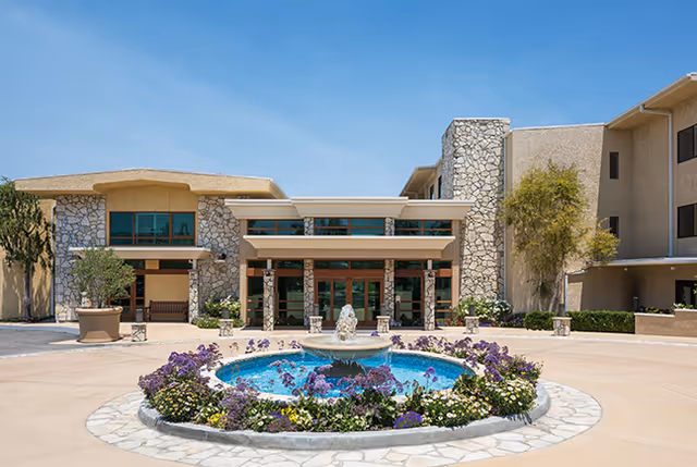 Front exterior view of The Canterbury Retirement Community building with a stone facade, large windows, and a covered entrance. In front of the building is a circular fountain surrounded by colorful flowers and plants.