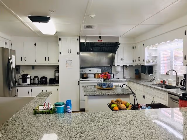 A bright, clean kitchen with white cabinets and granite countertops. The kitchen features a stainless steel refrigerator, a stove with a black range hood, a sink under a window with white blinds, and various kitchen appliances and utensils. There is a small island in the center with a decorative tray holding flowers and a bowl of fruit.