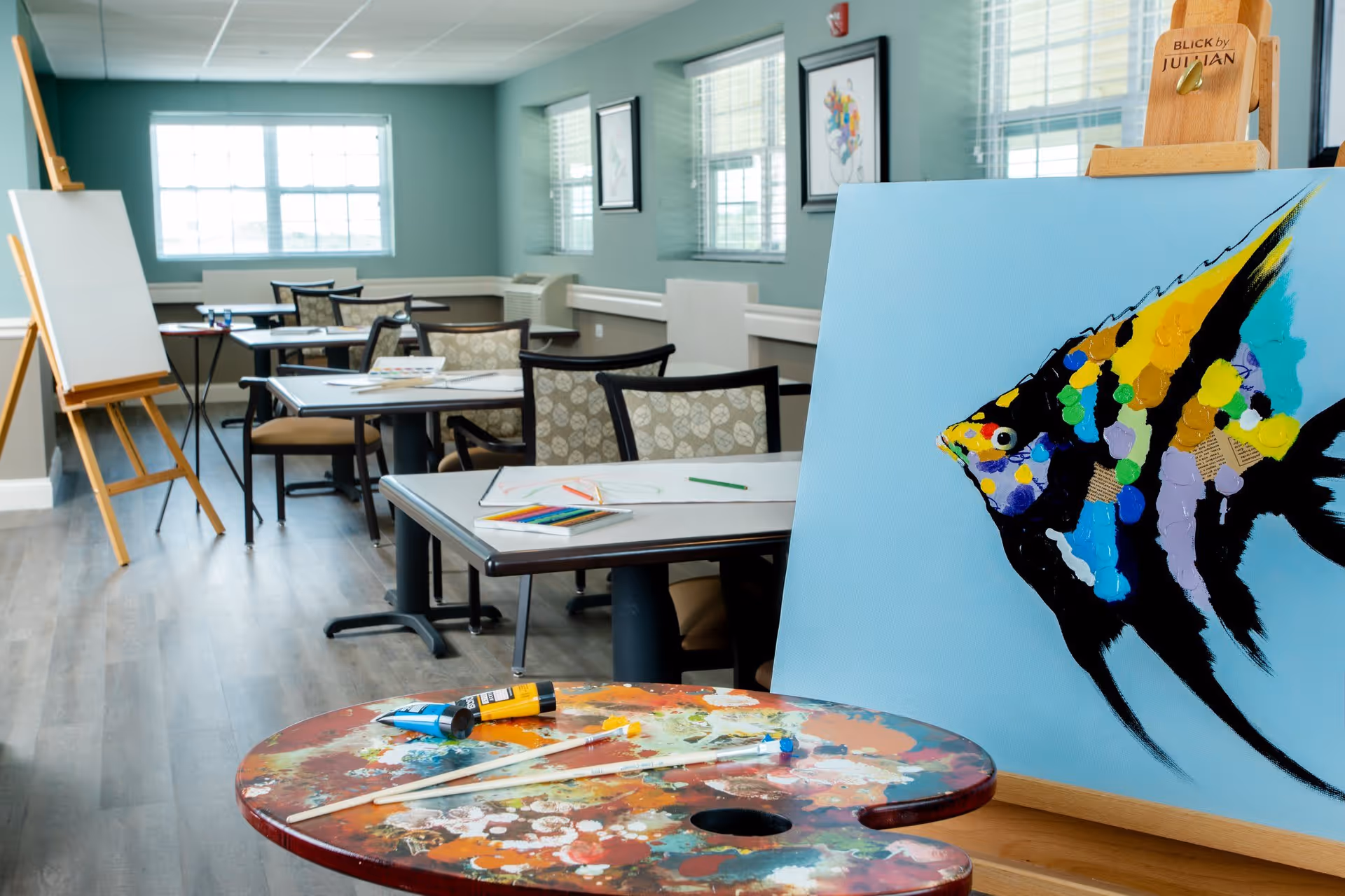 A bright art room with tables and chairs arranged for painting activities. In the foreground, there is a colorful painting of a fish on an easel and a paint palette with brushes and paint tubes. The room has large windows letting in natural light and framed artwork on the walls.