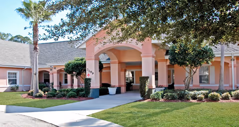 Front entrance of a pink single-story senior living building with a covered porte-cochère, columns, palm trees, and manicured landscaping.