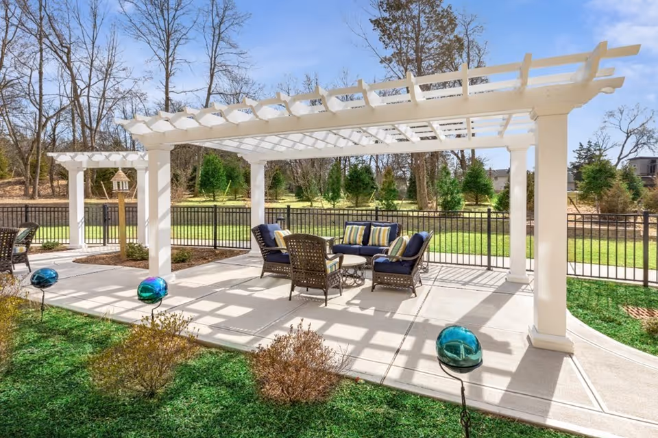 Outdoor patio area with white pergolas providing partial shade over a seating arrangement consisting of cushioned chairs and a small table. The patio is surrounded by a black metal fence, with green grass and small bushes in the foreground and trees in the background under a clear blue sky.
