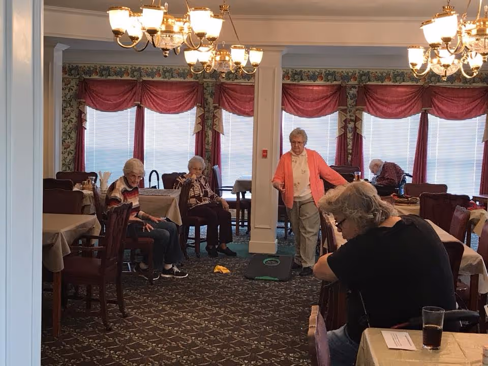 Several elderly residents sitting and socializing in a dining/activity room with tables, chandeliers, and red window valances.