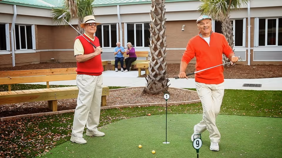Two elderly men playing mini golf on an outdoor putting green at a senior living facility. One man is wearing a red vest and hat, holding a golf club, while the other man in a red sweater is swinging a putter. In the background, two elderly people are sitting on a bench clapping and watching. The setting includes palm trees and a building with windows.