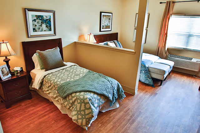 Bright furnished bedroom with a single bed, nightstand, and a small seating area by a window.