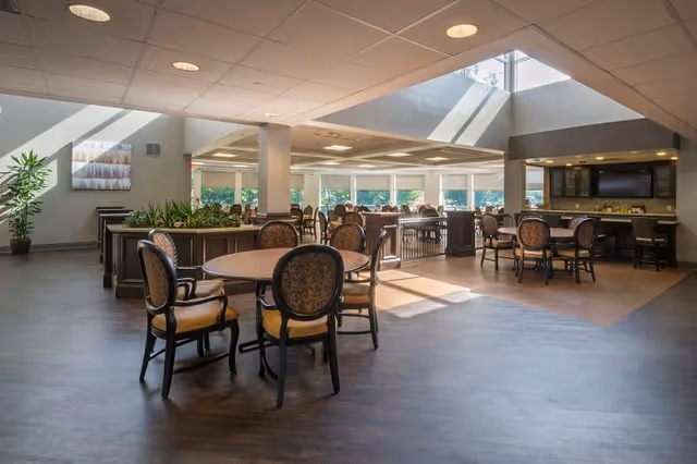 A spacious, well-lit dining area with multiple round tables and cushioned chairs arranged neatly. Large windows allow natural light to fill the room, and there is a skylight above. The room features a modern design with plants and a television mounted on the wall in the background.