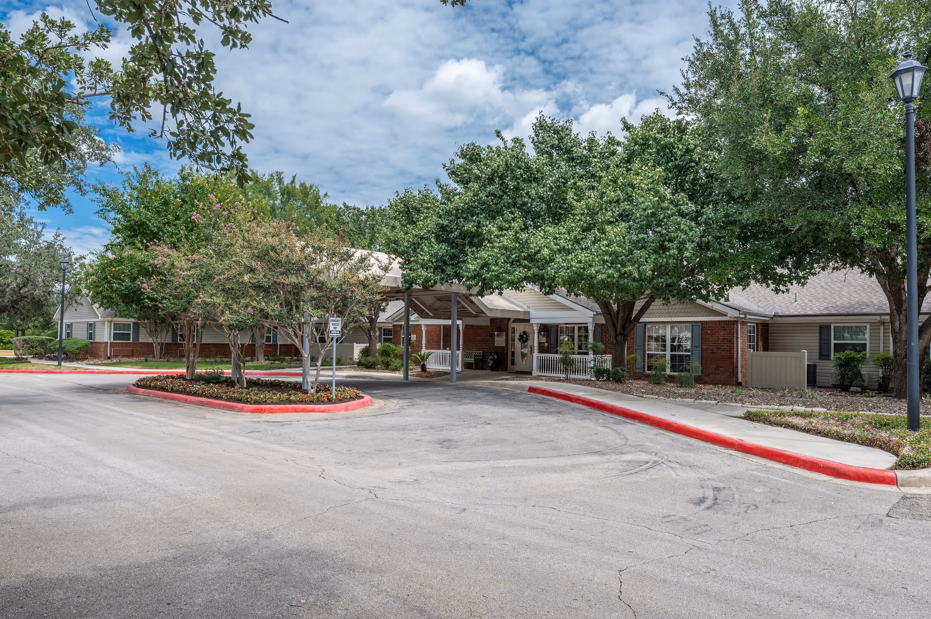 Exterior view of a senior living facility with a covered entrance, surrounded by trees and landscaping. The building has brick and siding walls with windows and a porch area. The driveway curves around a landscaped island with small trees and flowers.