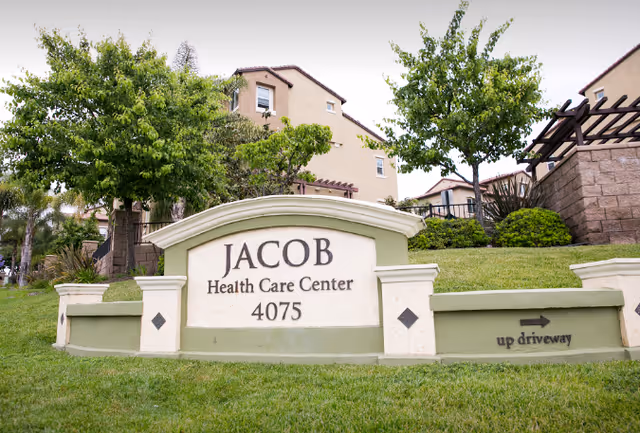 Outdoor view of the entrance sign for Jacob Health Care Center with the address 4075, surrounded by green grass, trees, and residential-style buildings in the background.