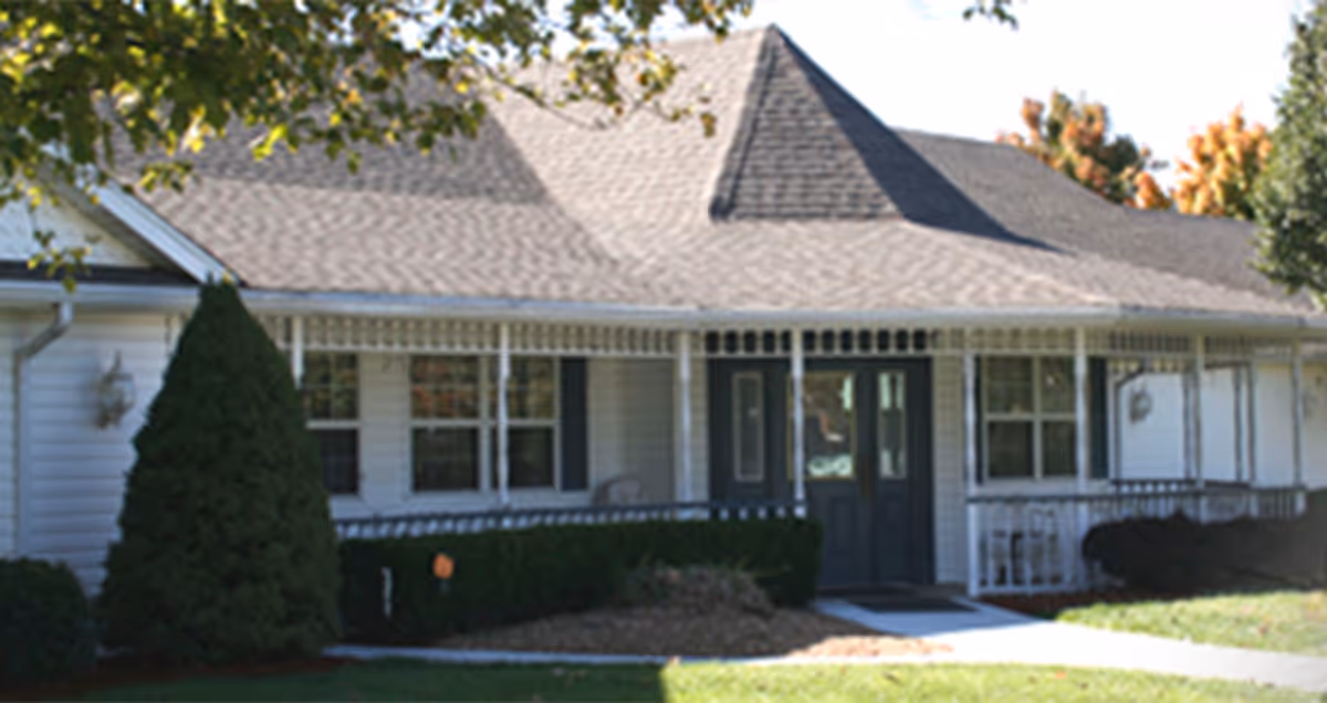 Exterior view of a single-story residential building with a gray shingled roof, white siding, and a covered front porch. There are several windows with black shutters and a double front door. The building is surrounded by green bushes, a tree with green leaves, and a well-maintained lawn.