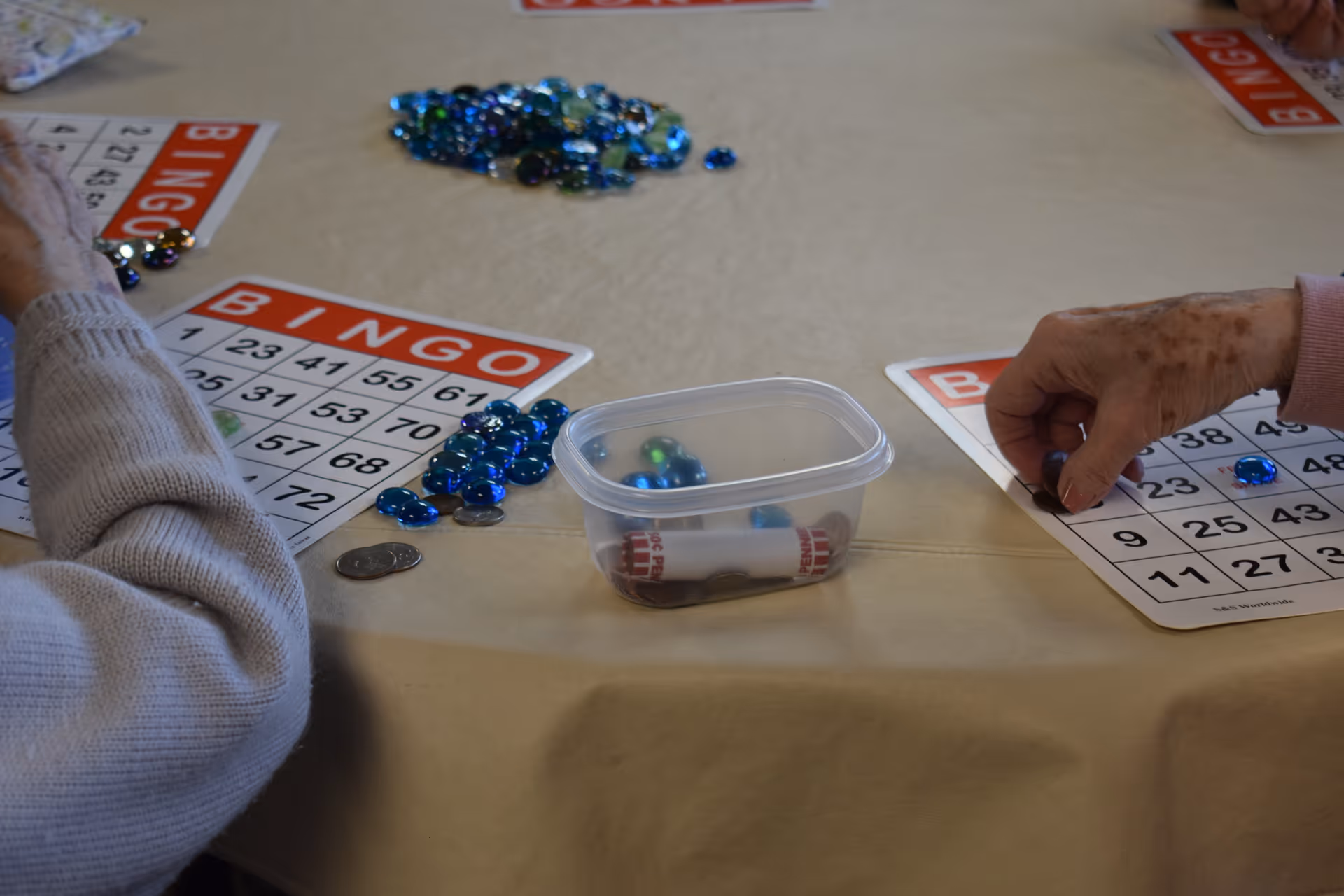 Two elderly individuals playing bingo at a table covered with a beige tablecloth. Each person has a bingo card in front of them, and there are blue and clear glass markers placed on the cards. A small plastic container with additional markers and coins is on the table.