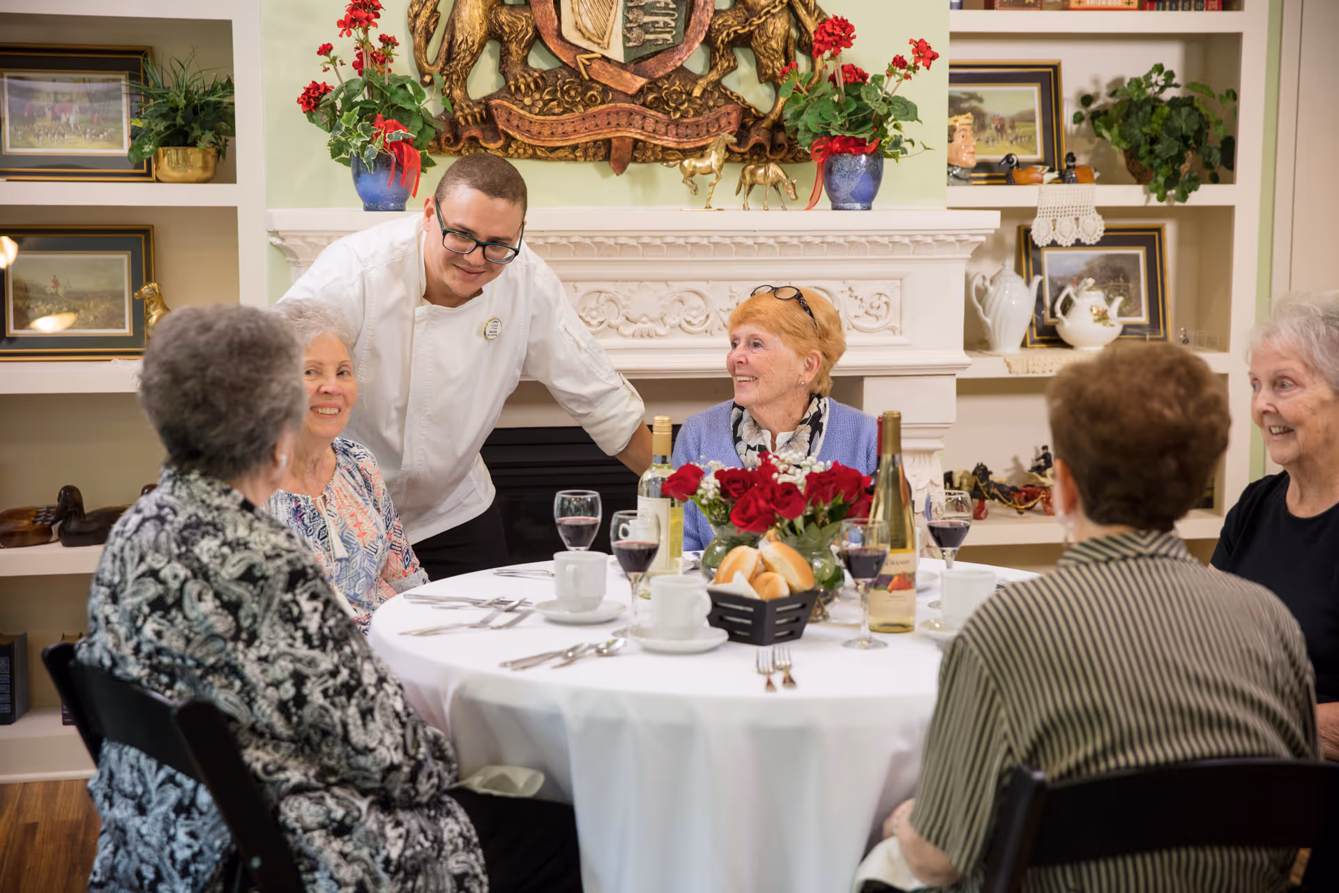 A group of five elderly women seated around a round dining table covered with a white tablecloth, engaged in conversation with a smiling male staff member leaning in to talk with them. The table is set with wine glasses, coffee cups, plates, cutlery, a basket of bread rolls, and a vase of red roses. The background features a decorative white fireplace mantel with ornate carvings, red flowers in blue vases, and shelves with framed pictures and decorative items.