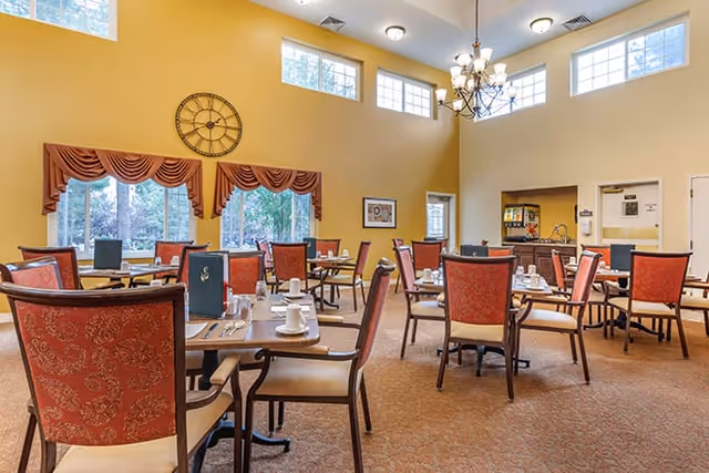A bright dining room with several tables and red cushioned chairs arranged neatly. Each table is set with white cups, saucers, and silverware. Large windows with red valances allow natural light to fill the room. A large decorative clock hangs on the yellow wall, and a chandelier is suspended from the high ceiling.