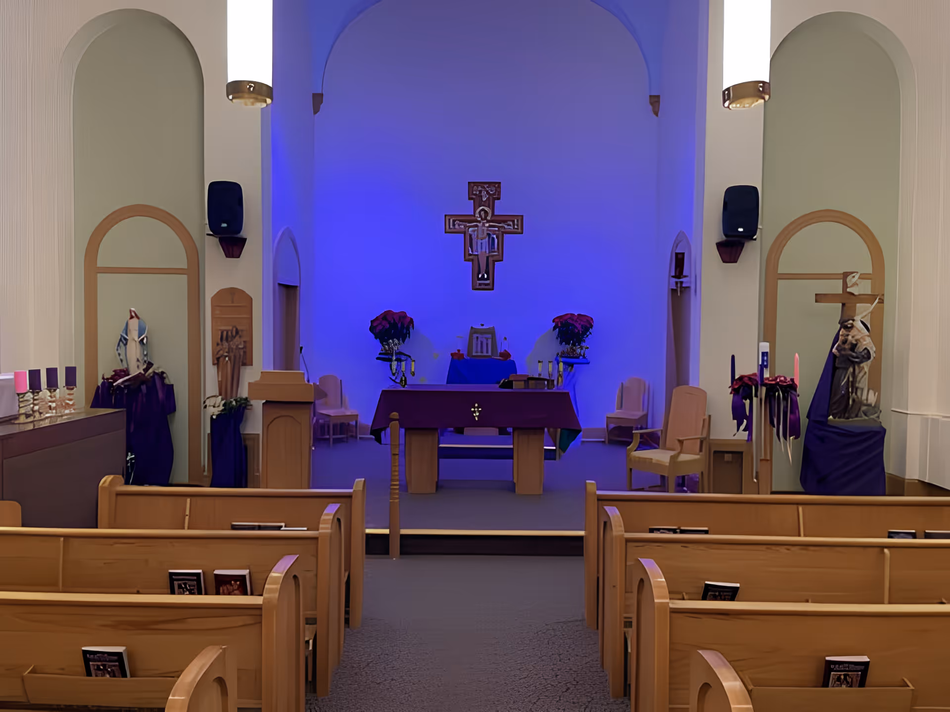 Interior view of a chapel with wooden pews facing an altar. The altar is decorated with purple cloth and flowers, and a crucifix is mounted on the wall behind it. The chapel has arched alcoves on either side, some with religious statues and candles. The lighting casts a blue hue on the altar area.