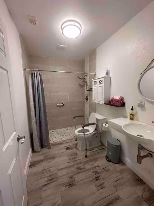 A senior-friendly bathroom with a walk-in shower featuring a curtain and grab bar, a toilet with safety rails, a wall-mounted sink, a small cabinet above the toilet, a trash can, and a round mirror on the wall.