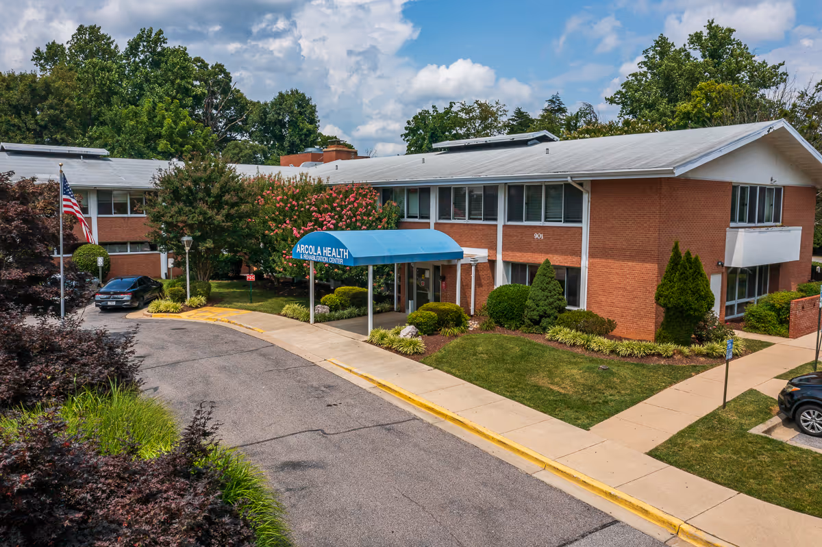 Two-story brick senior healthcare building with a blue entry awning reading 'Arcola Health', landscaped grounds and a curved driveway.