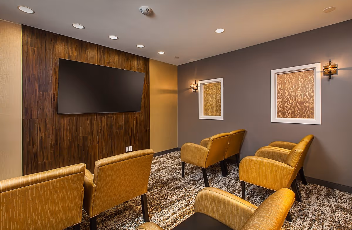 A small media room with six mustard yellow upholstered chairs arranged in two rows facing a large flat-screen TV mounted on a dark wood-paneled wall. The room has a patterned carpet, gray and beige walls, two framed decorative panels, and wall sconces providing warm lighting.