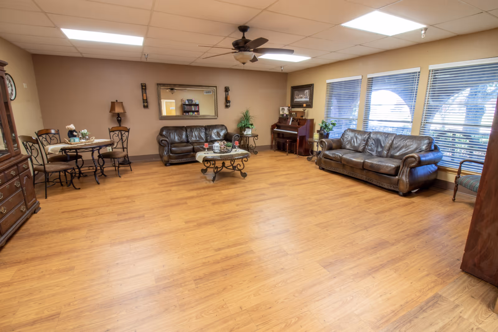 Spacious senior living common room with leather sofas, a coffee table, a small dining set, and a piano on hardwood floors by large windows.