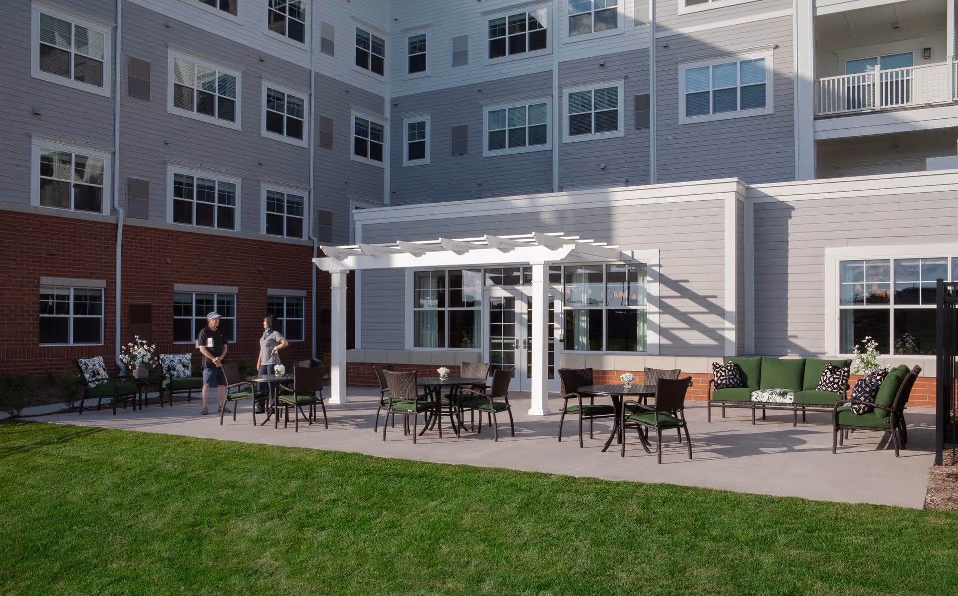 Outdoor patio area at Clarendale of Addison with green cushioned chairs and sofas, round tables, a white pergola, and two people standing and talking near the building. The building has multiple windows and a combination of gray siding and red brick exterior.