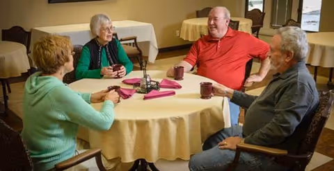 Four elderly people sitting around a round table covered with a beige tablecloth, each holding a coffee mug and engaging in conversation in a common area with additional tables and chairs in the background.