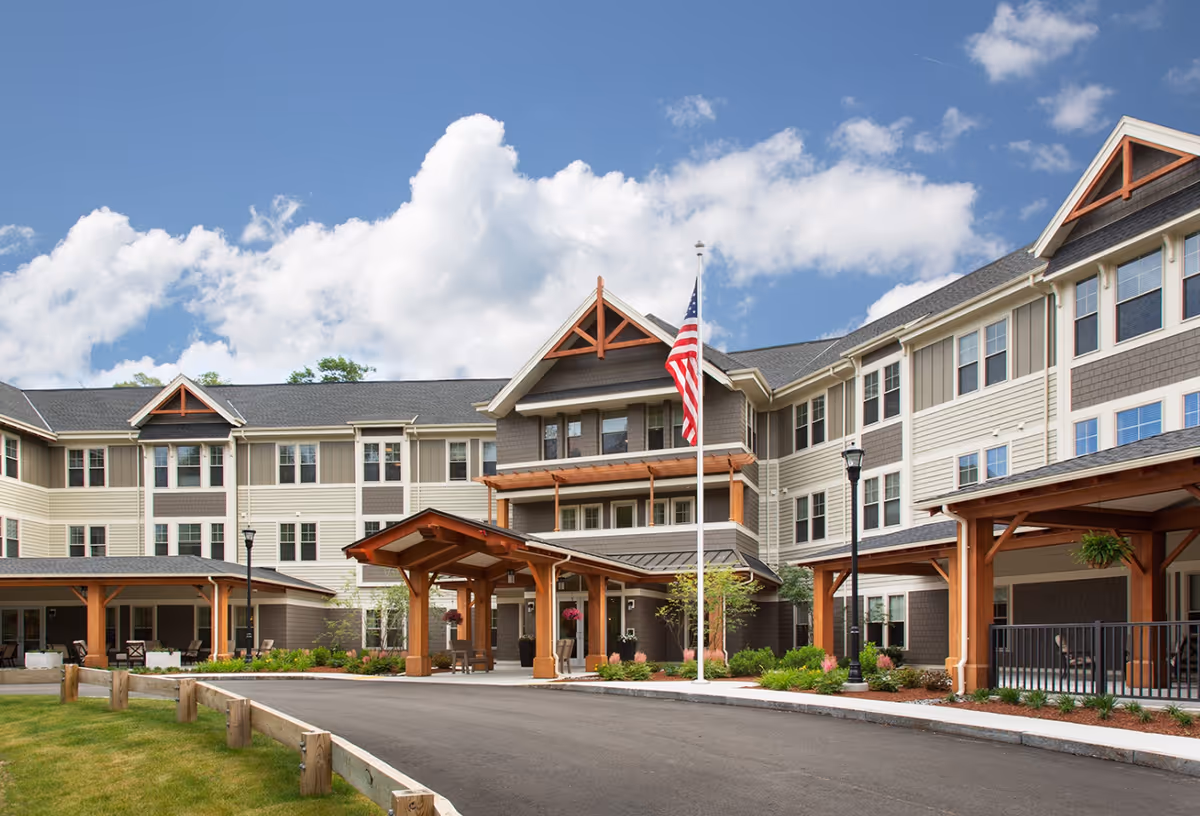 Front exterior of a three-story senior living building with a covered entrance, American flag, and landscaped driveway.