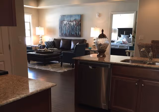 Open-concept kitchen and living room with a granite-topped island and sink in the foreground and a leather sofa, armchairs, and lamps in the living area with a bedroom visible through a doorway.