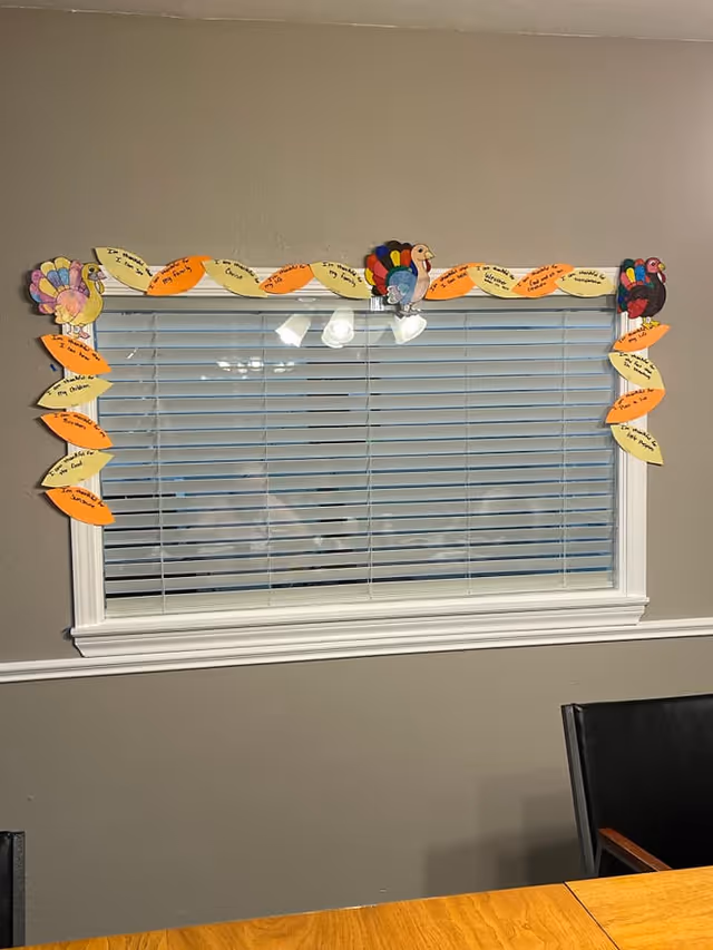 A window with closed white blinds in a room with gray walls. The window frame is decorated with colorful paper turkeys and orange and yellow paper leaves, each leaf containing handwritten messages. Below the window is a wooden table and a black chair with wooden armrests.