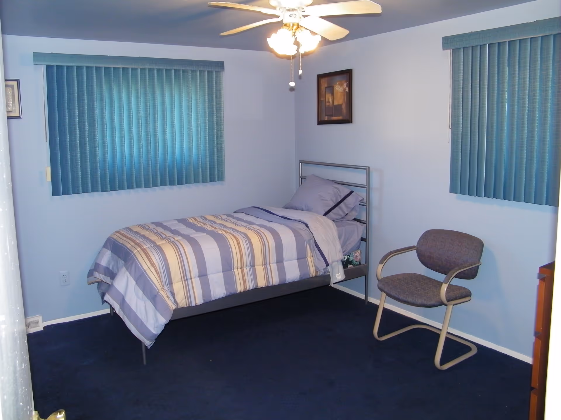 Simple bedroom with a twin bed dressed in striped bedding, a cushioned chair, two windows with teal vertical blinds and a ceiling fan.