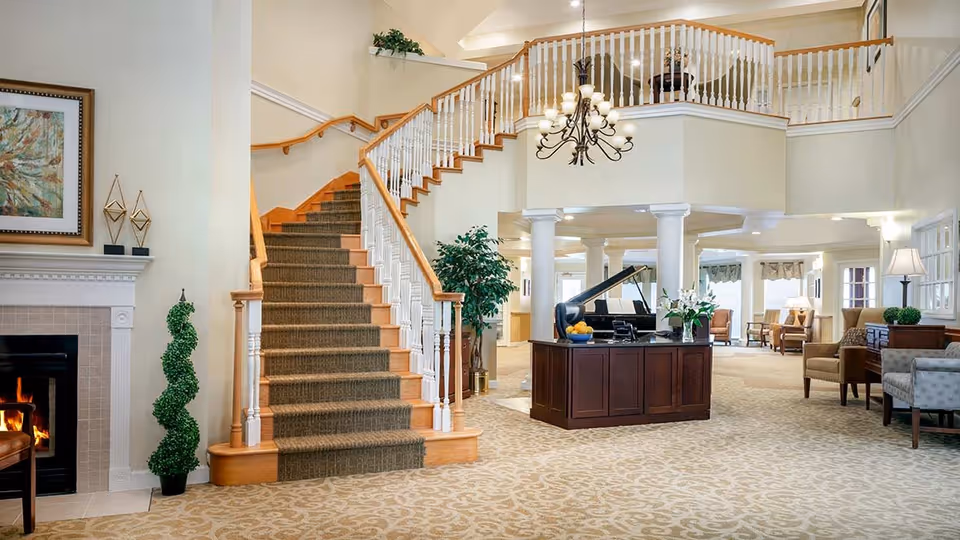 Spacious and elegant interior of a senior living facility featuring a carpeted staircase with wooden handrails, a chandelier hanging from the ceiling, a fireplace with a fire burning, a grand piano on a wooden desk, several armchairs, and decorative plants and artwork.