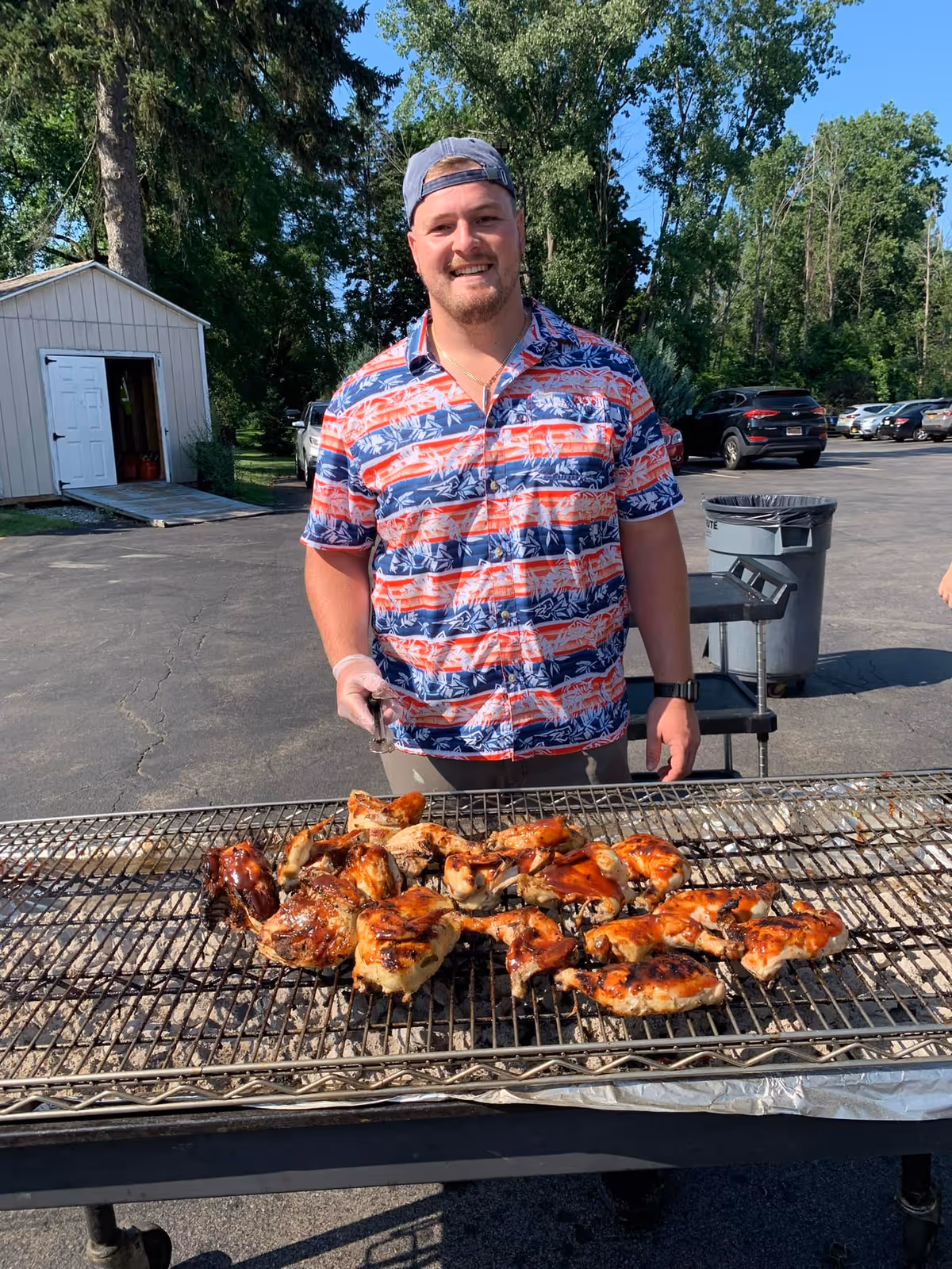 A man wearing a colorful patterned shirt and a backwards cap is standing outdoors behind a large grill with several pieces of grilled chicken. There are trees, parked cars, and a small shed in the background under a clear blue sky.