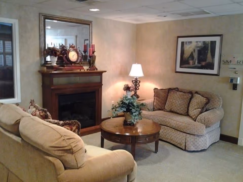 A cozy living room with two patterned sofas facing each other, a round wooden coffee table with a potted plant, a table lamp on a side table, a wooden fireplace with decorative items including a clock and candles, a large mirror above the fireplace, and a framed picture on the wall.