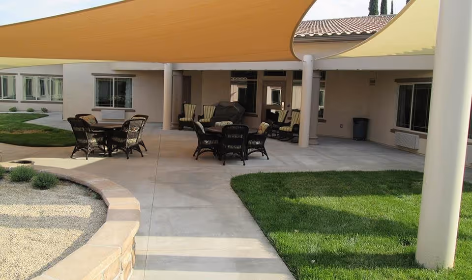 Outdoor patio area with tables and chairs under large shade sails, surrounded by grass and a building with windows and a tiled roof in the background.