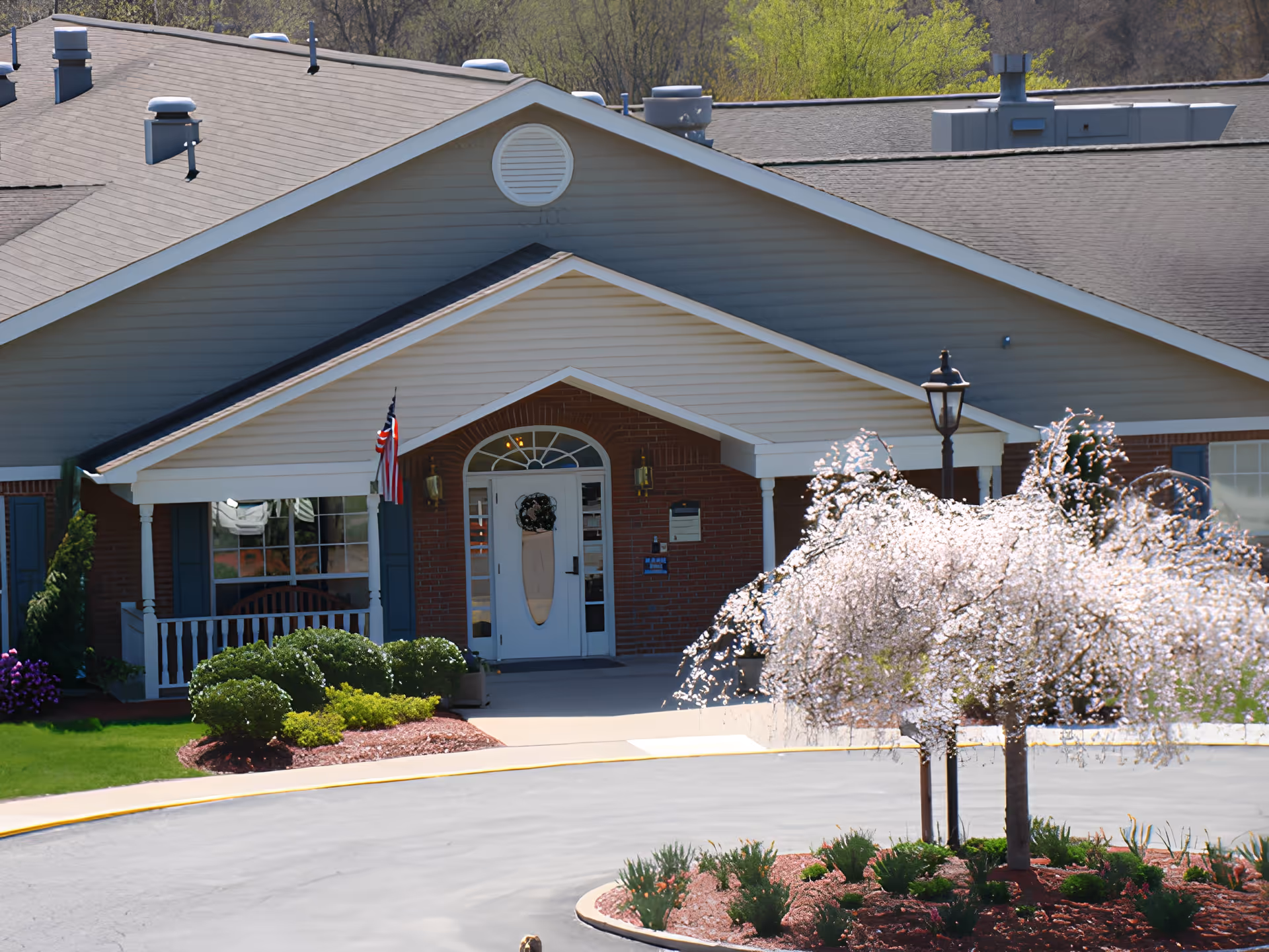 Front entrance of a brick-and-siding senior care facility with a covered porch, American flag, and a landscaped circular driveway with a blooming tree.