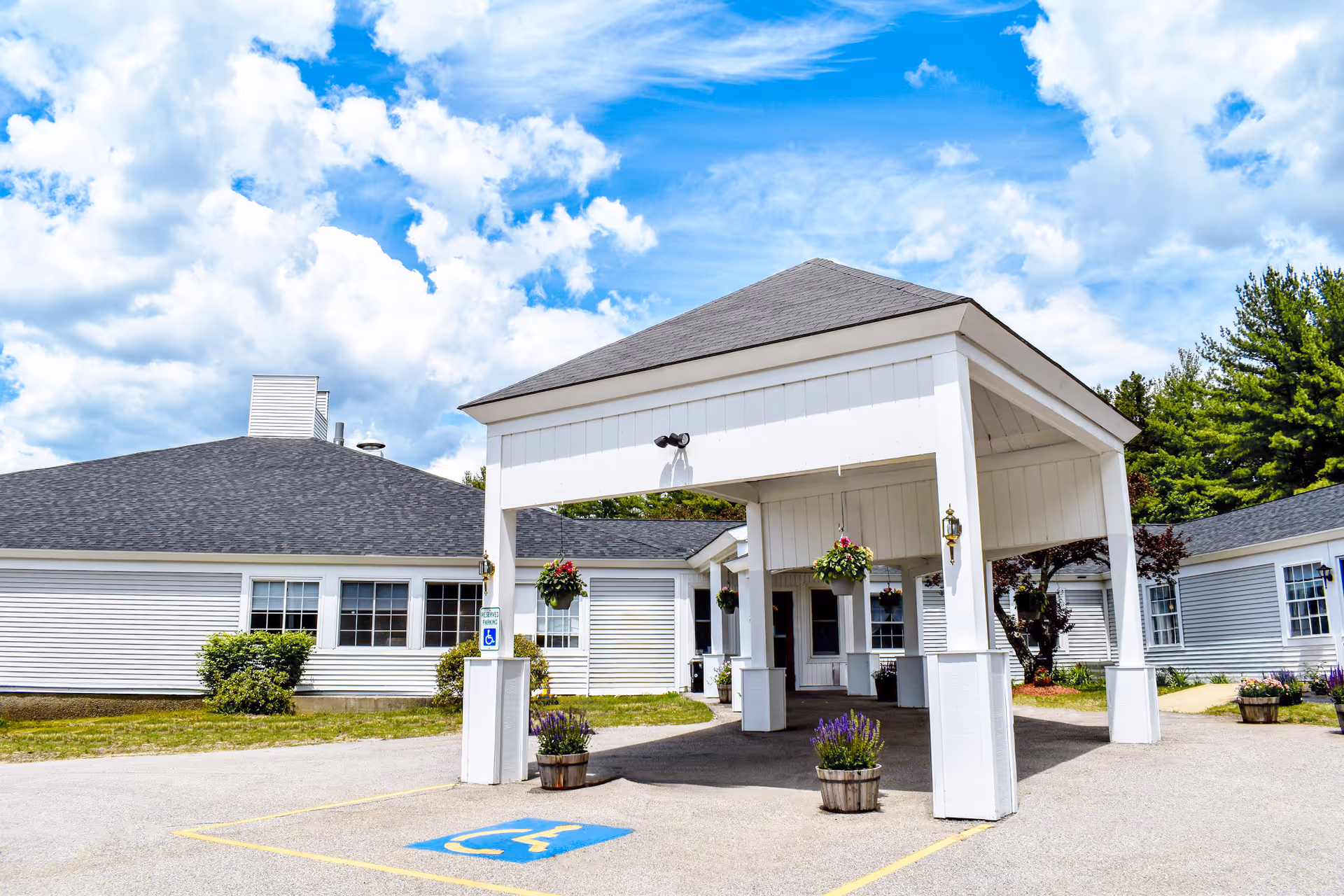 White single-story senior living building with a covered entrance canopy, hanging flower baskets, and a marked handicap parking space.
