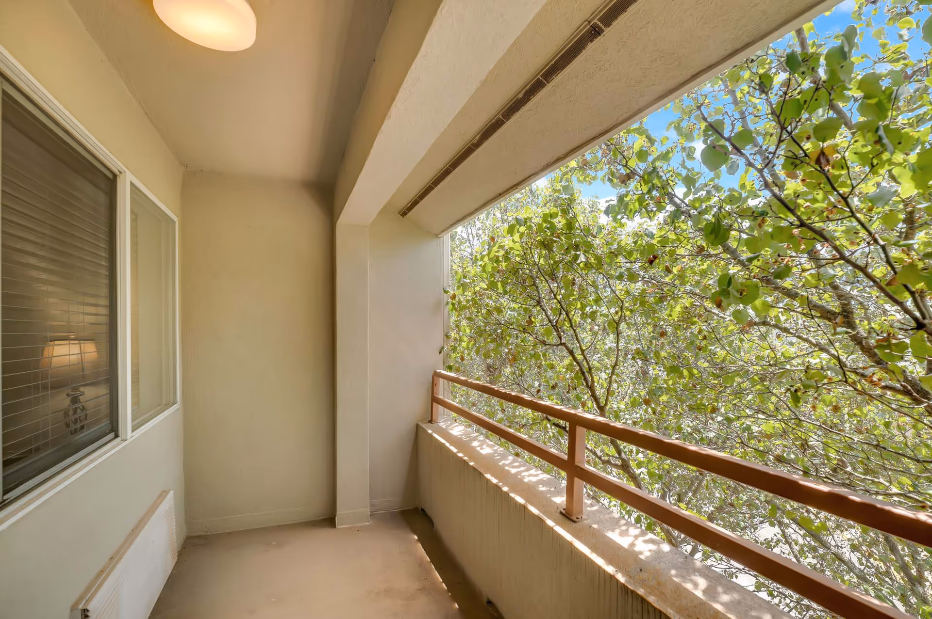 A small covered balcony with beige walls and floor, a brown railing, and a view of green leafy trees under a blue sky. A window with closed blinds and a lamp inside the adjacent room is visible on the left side.