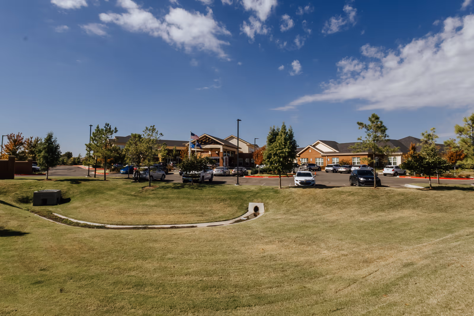 Exterior view of The Harrison of Oklahoma City Assisted Living & Memory Care facility with a parking lot, several cars, trees, and a grassy area under a partly cloudy blue sky.