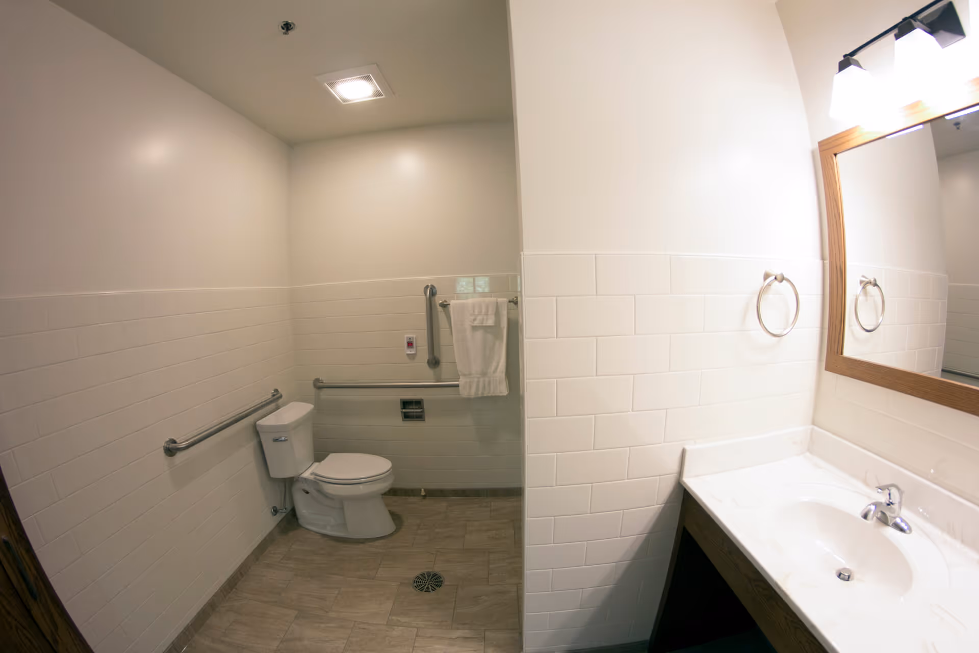 A clean and accessible bathroom with white tiled walls and beige floor tiles. The room features a toilet with grab bars on both sides, a towel hanging on a bar, a sink with a countertop, a mirror with a wooden frame, and a light fixture above the mirror.