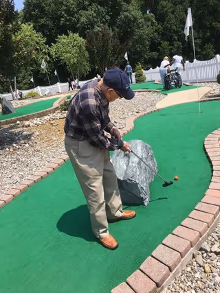 An elderly man wearing a plaid shirt, beige pants, and a navy cap is playing mini golf on a green artificial turf course bordered by bricks and rocks. In the background, there are other people, including one in a wheelchair, near a white picket fence and trees.