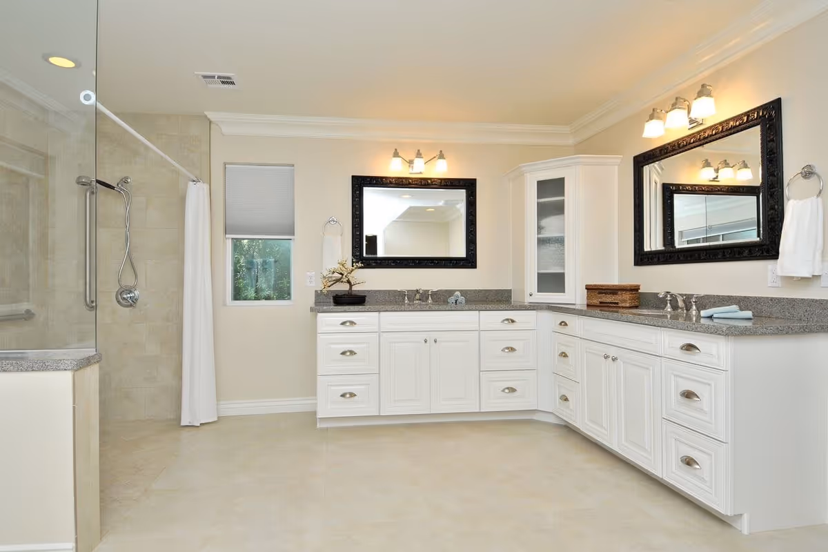 A spacious, well-lit bathroom featuring a large vanity with white cabinets and granite countertops, two sinks with chrome faucets, two large framed mirrors above each sink, a glass-enclosed shower with a handheld showerhead, a small window with a closed blind, and beige tiled floors and walls.