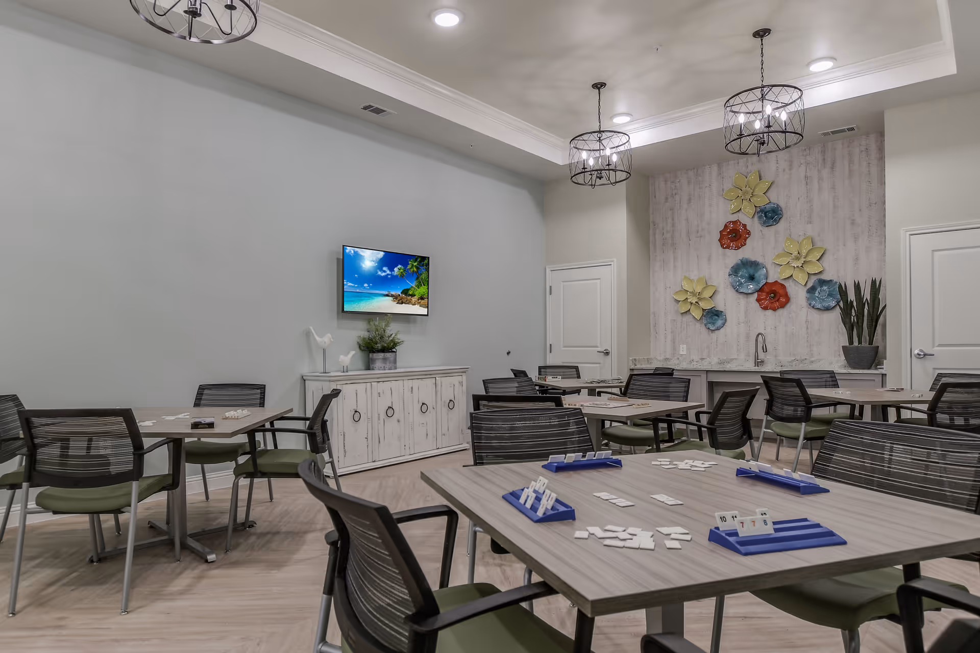 A well-lit activity room with multiple tables and chairs arranged for playing games. The tables have game tiles and racks on them. The room features a wall-mounted TV displaying a beach scene, decorative wall art with colorful flowers, and two modern pendant lights hanging from the ceiling. There is a white cabinet under the TV and a small sink area with a plant on the counter.