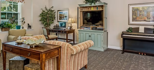 A cozy living room area with a striped sofa facing a green entertainment center holding a flat-screen TV. There is a wooden table behind the sofa with decorative items, a piano against the wall, a desk with a lamp and chair, and framed artwork on the walls. A window with plants on the sill lets in natural light.