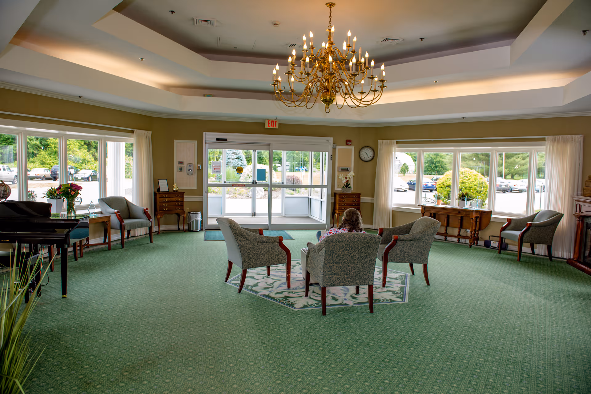 A spacious, well-lit common area with green carpet and a chandelier hanging from the ceiling. Four upholstered chairs are arranged around a small rug in the center, with a person sitting in one of the chairs. Large windows let in natural light and offer views of the parking lot and greenery outside. There is a piano on the left side and wooden furniture along the walls.